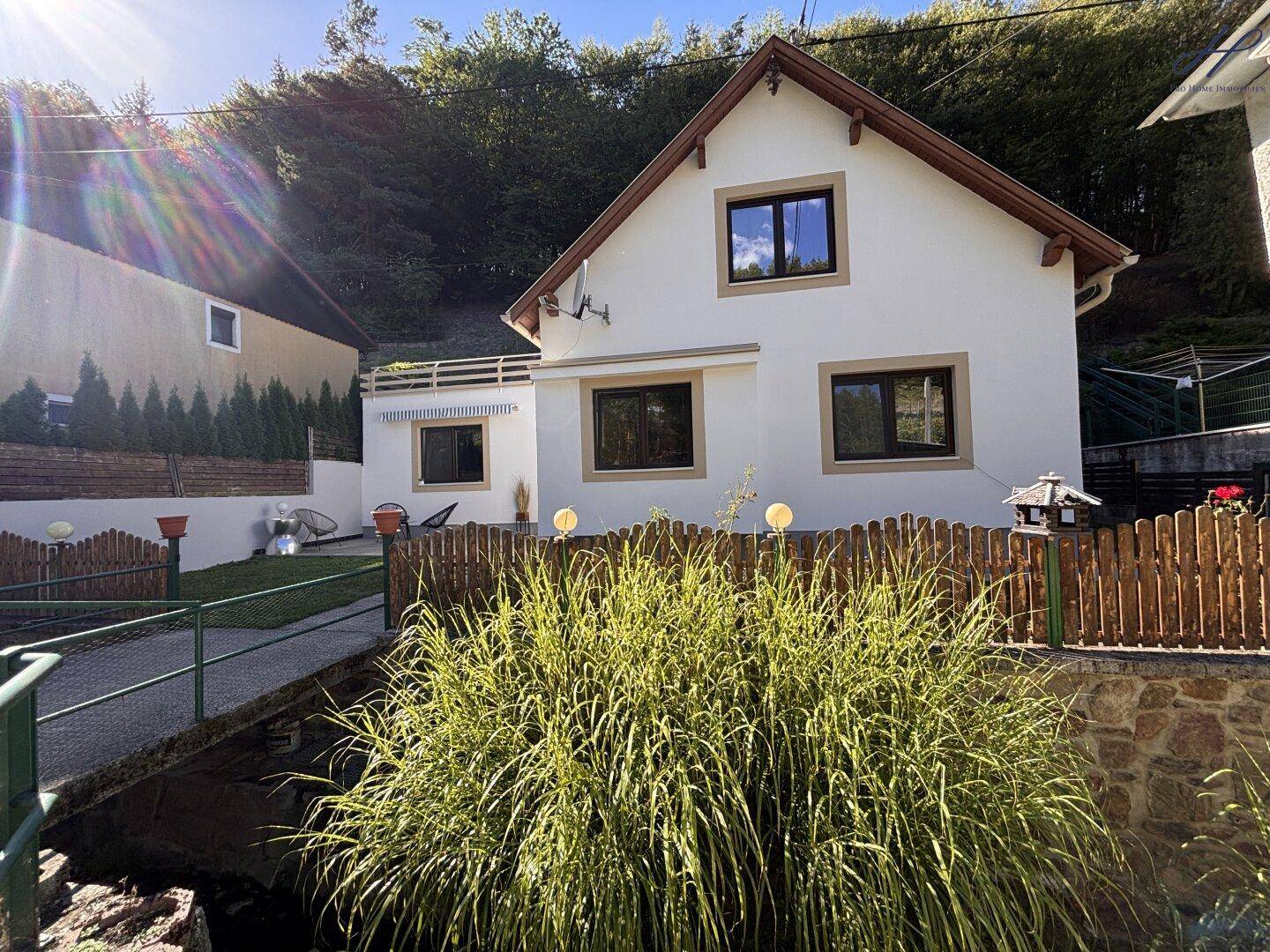 Sonniger Blick auf das weiße Einfamilienhaus mit braunen Fenstern und bewaldeter Umgebung.
