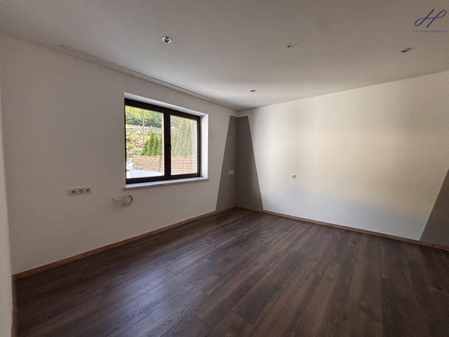 Leerer Raum mit Laminatboden, weißen Wänden und einem Fenster mit Blick ins Grüne.