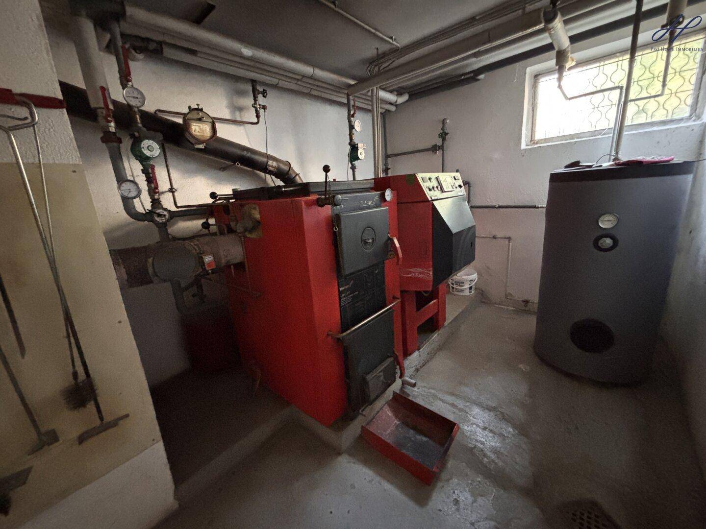 Funktionaler Heizungsraum im Keller mit einem großen roten Heizkessel, Warmwasserspeicher und sichtbaren Rohrleitungen.