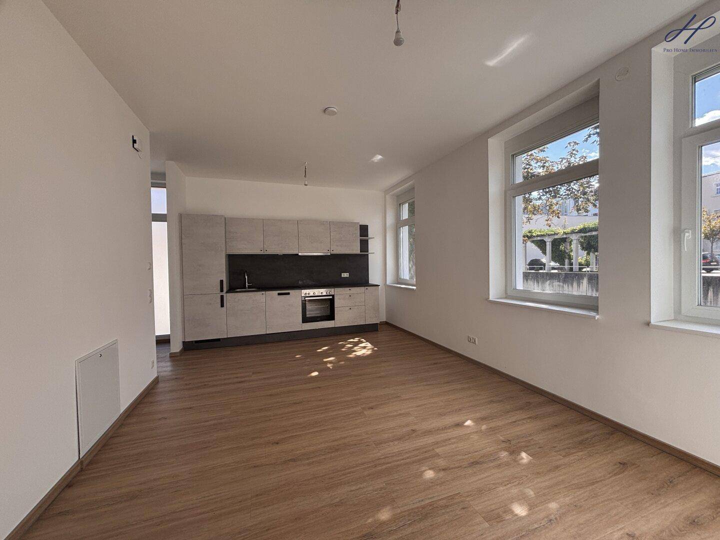 Geräumiger Wohnbereich mit Küchenzeile, Laminatboden und viel Tageslicht durch große Fenster.