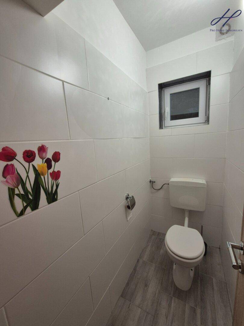 Kleines WC mit weißen Fliesen, Toilette und einem Fenster, verziert mit floralen Akzenten.