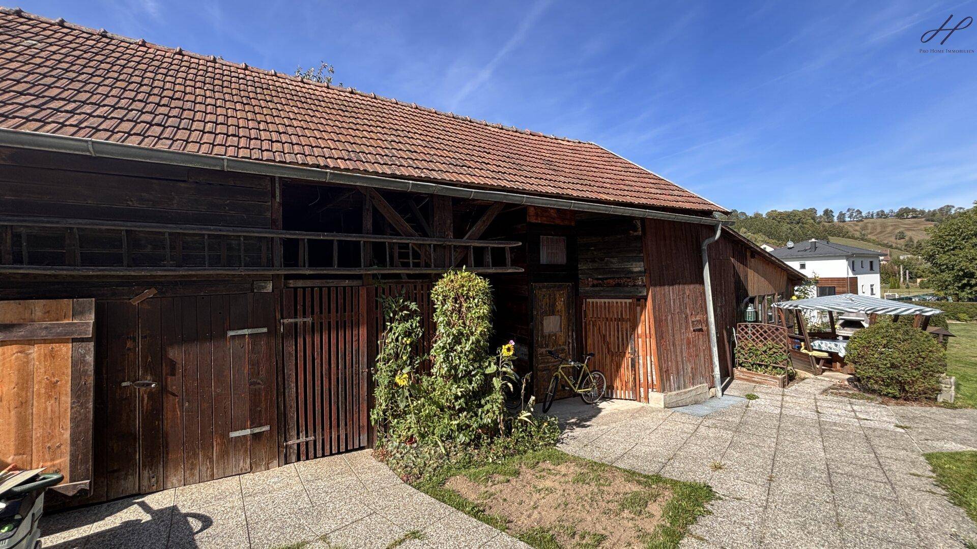Großes hölzernes Nebengebäude mit Ziegeldach und gepflastertem Vorplatz, umgeben von Natur.