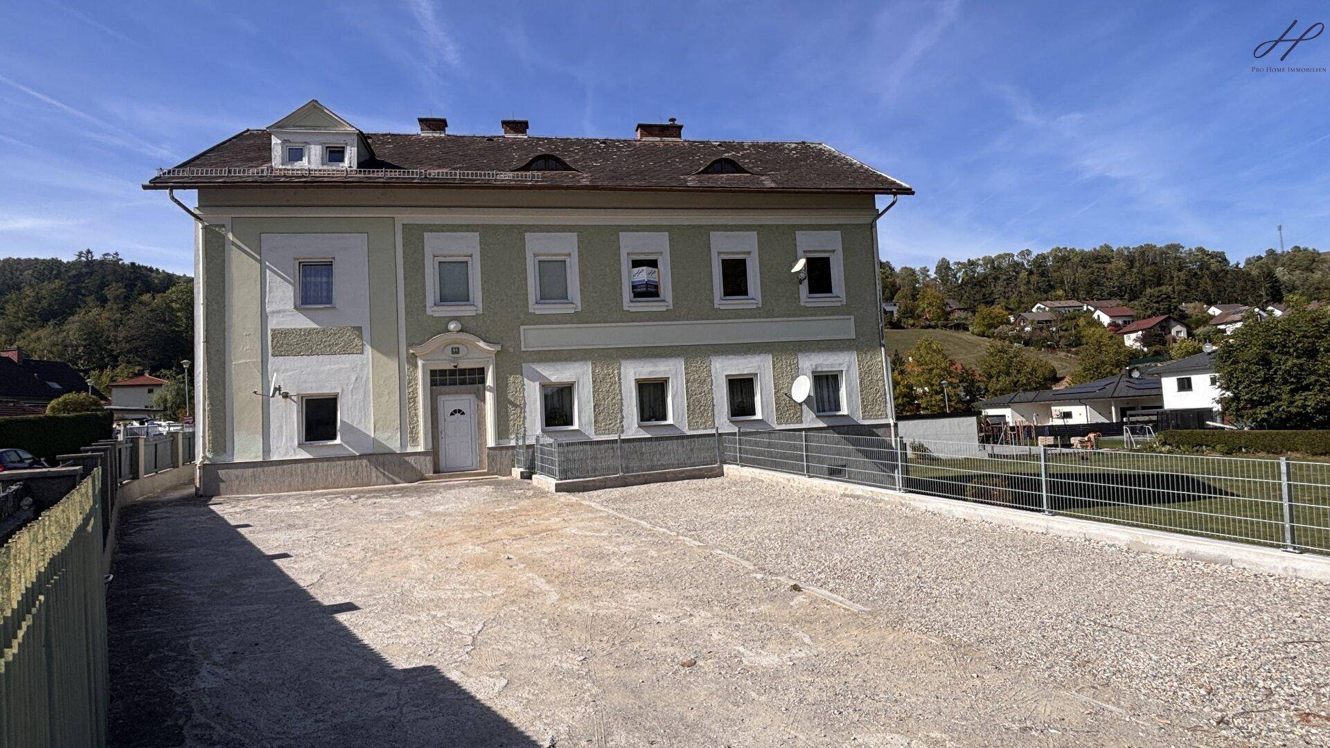 Vorderansicht des Hauses mit heller Fassade, Schotterauffahrt und Blick auf die umliegende Hügellandschaft.