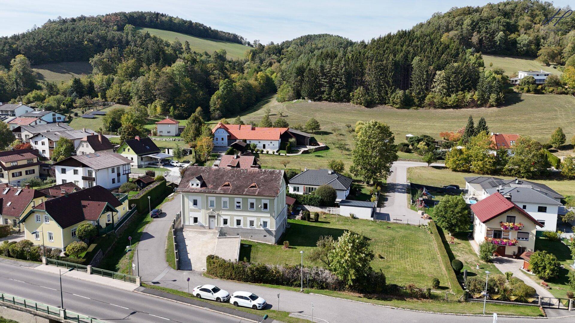Luftaufnahme eines weitläufigen Grundstücks mit einem historischen Gebäude und Blick auf die bewaldeten Hügel.