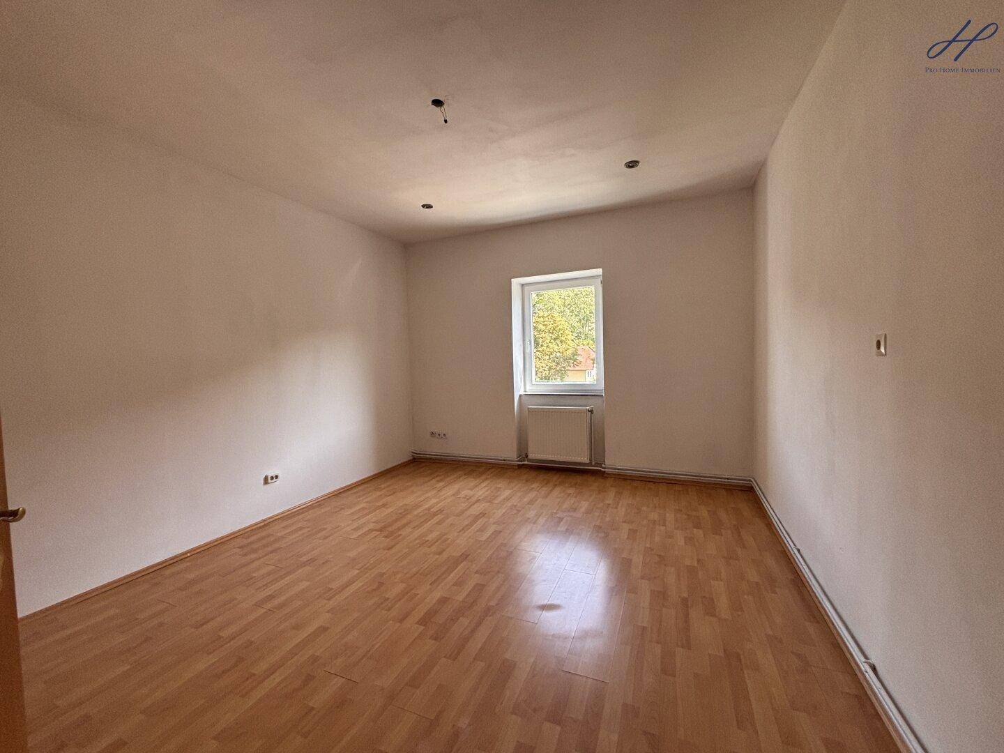 Leerer Raum mit Laminatboden und einem Fenster, das den Blick ins Grüne freigibt.