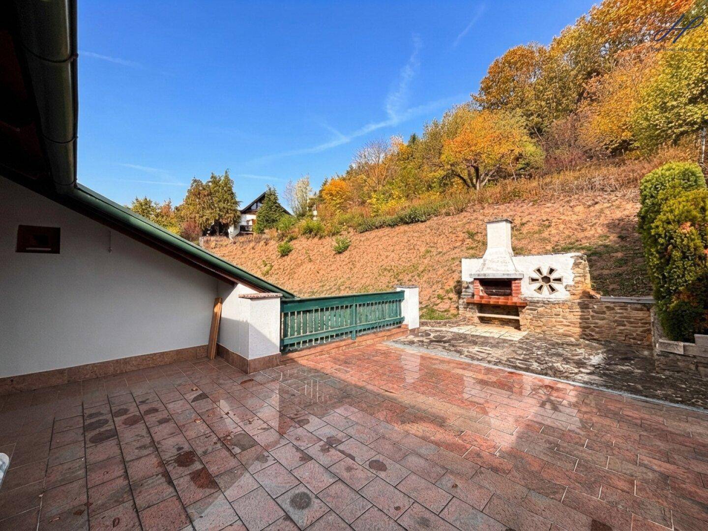 Große geflieste Terrasse mit gemauertem Grillplatz und Blick auf den bewaldeten Hang.