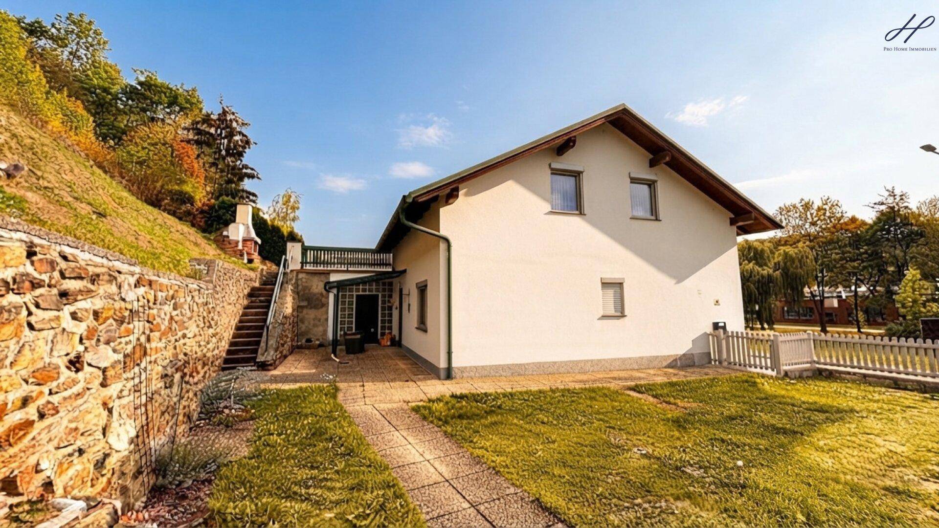 Haus mit weißer Fassade, grünem Rasen und Steintreppe, umgeben von Bäumen und Natur.
