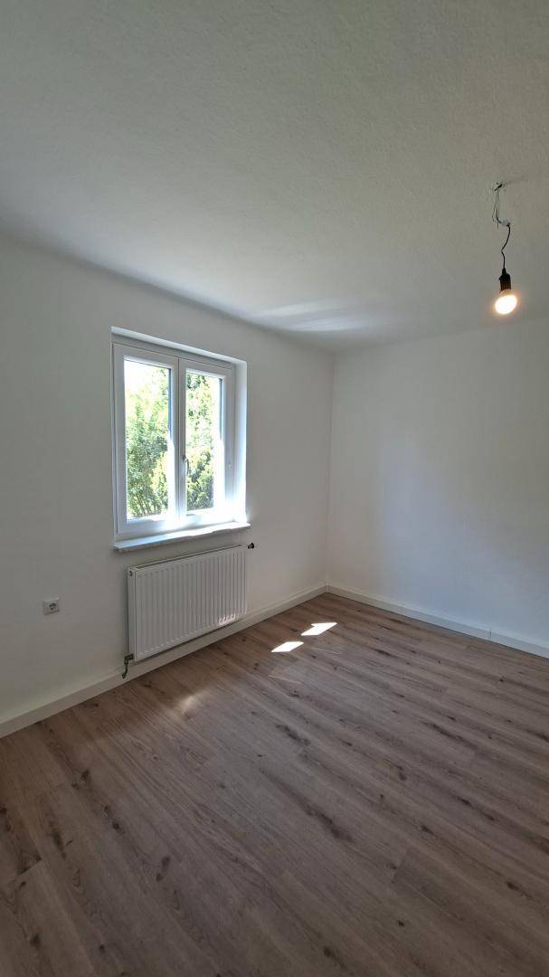 Leeres Zimmer mit Laminatboden, weißen Wänden und großem Fenster für viel Tageslicht.