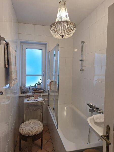 Helles Badezimmer mit Badewanne, Duschabtrennung und Fenster, beleuchtet von einem Kronleuchter.