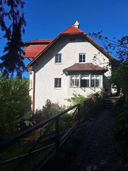 Traditionelles Haus mit rotem Dach und Erkerfenster, umgeben von üppigem Grün und einem Weg.