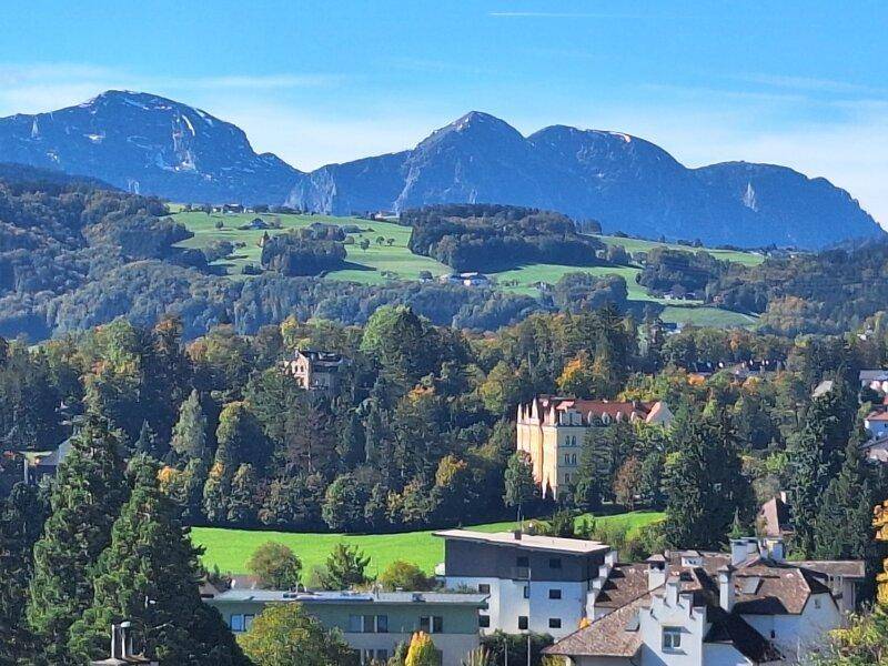 Panoramablick auf majestätische Berge, grüne Hügel und bewaldete Täler mit vereinzelten Gebäuden in der Ferne.