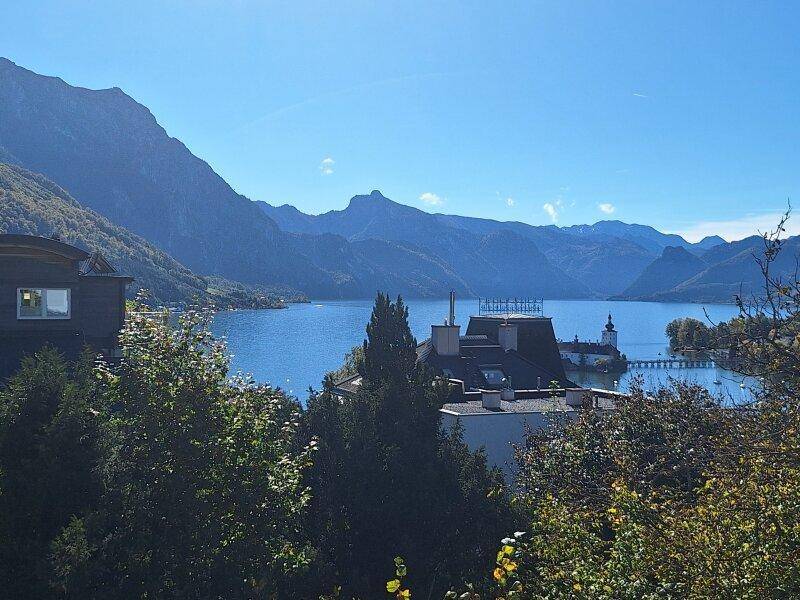 Weitläufiger Panoramablick auf den See und die umliegende Berglandschaft mit einem Schloss im Hintergrund.
