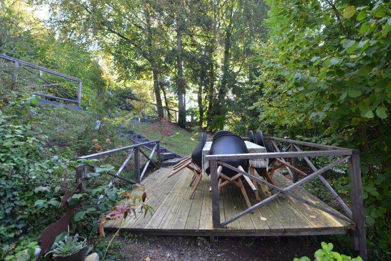 Holzterrasse mit Esstisch und Stühlen, umgeben von üppigem Grün und Bäumen.