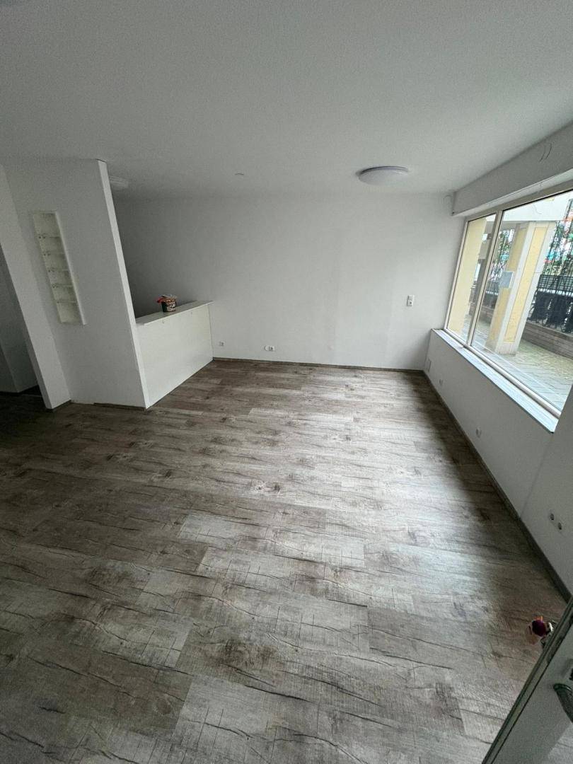 Heller, leerer Raum mit Laminatboden und großen Fenstern, die den Blick nach draußen ermöglichen.
