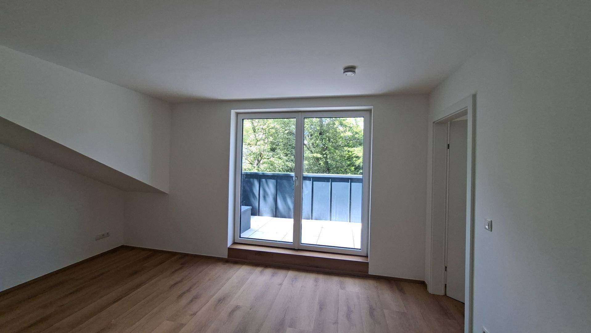 Heller Raum mit Holzboden, Dachschräge und Zugang zu einem Balkon mit grüner Aussicht.
