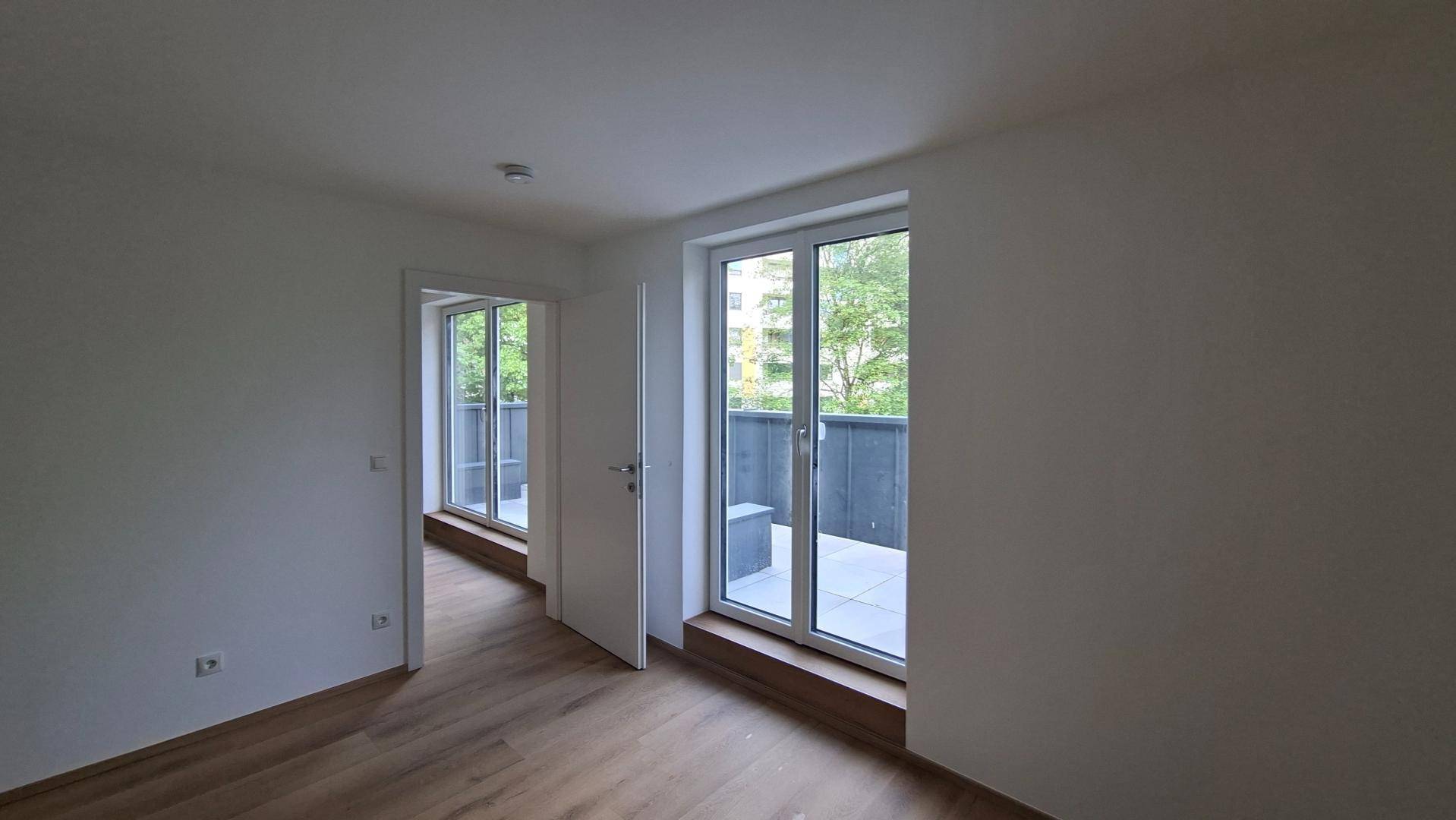 Lichtdurchfluteter Raum mit Holzboden und zwei Zugängen zu Außenbereichen mit grüner Umgebung.