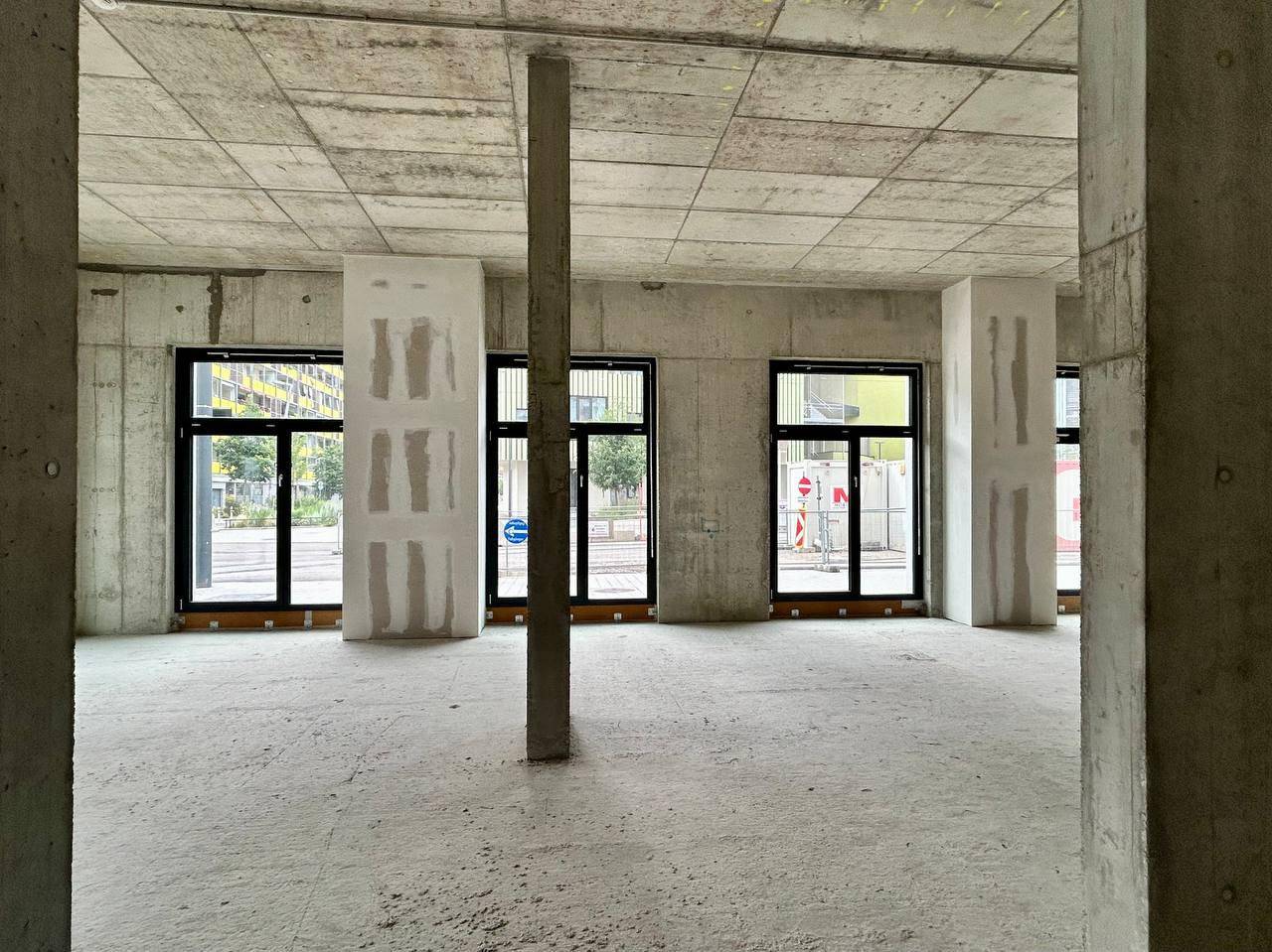 Unfertiger Innenraum mit Betonwänden, Säulen und großen Fensteröffnungen im Rohbauzustand.