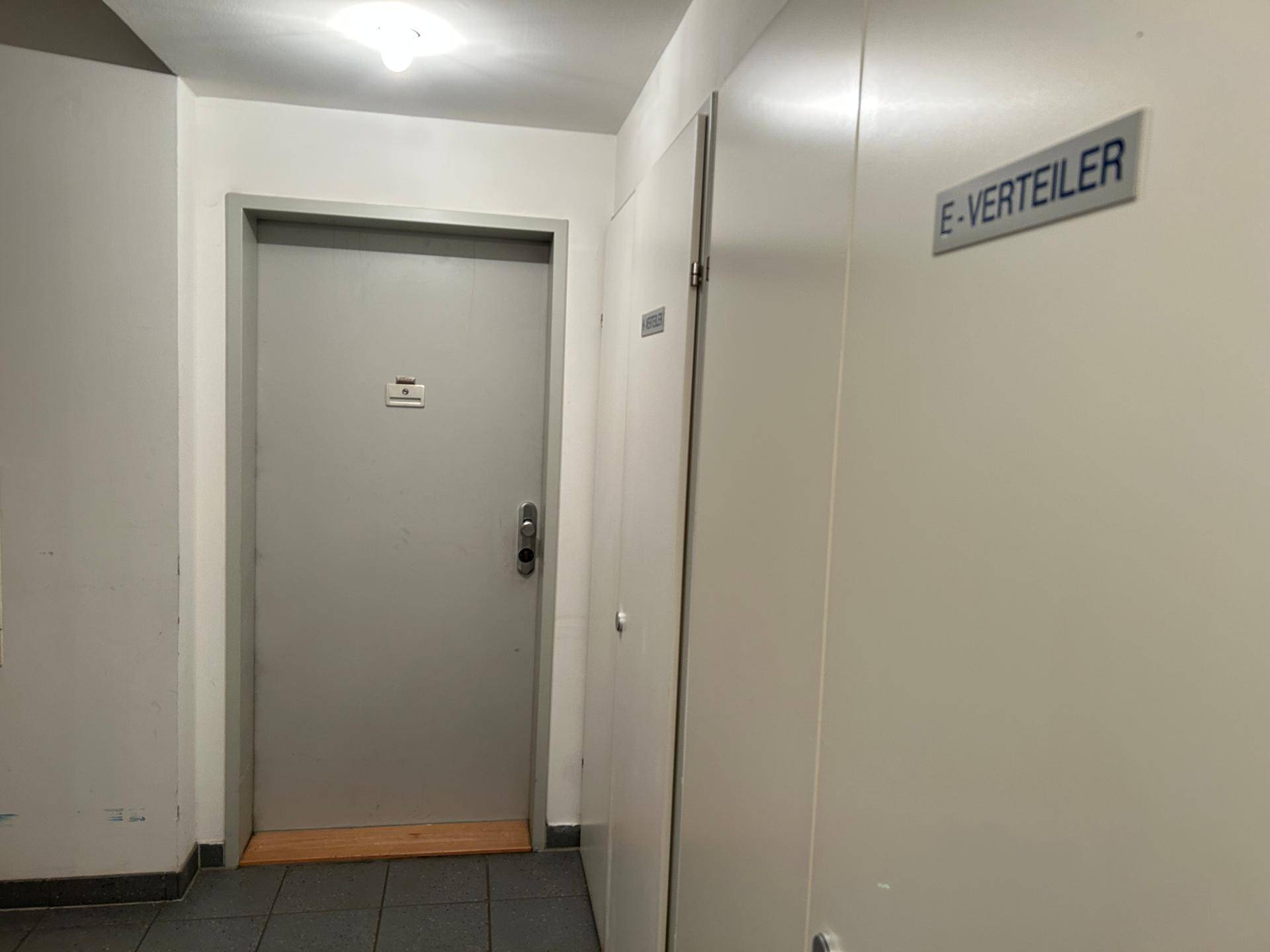 Funktionaler Flur mit grauer Tür und einem beschrifteten Elektroschrank, schlicht gestaltet.