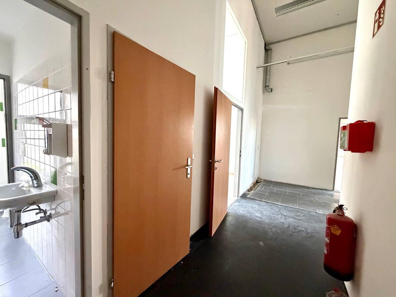 Flur mit Zugang zu Toilettenräumen, dunklem Boden und einem roten Feuerlöscher.