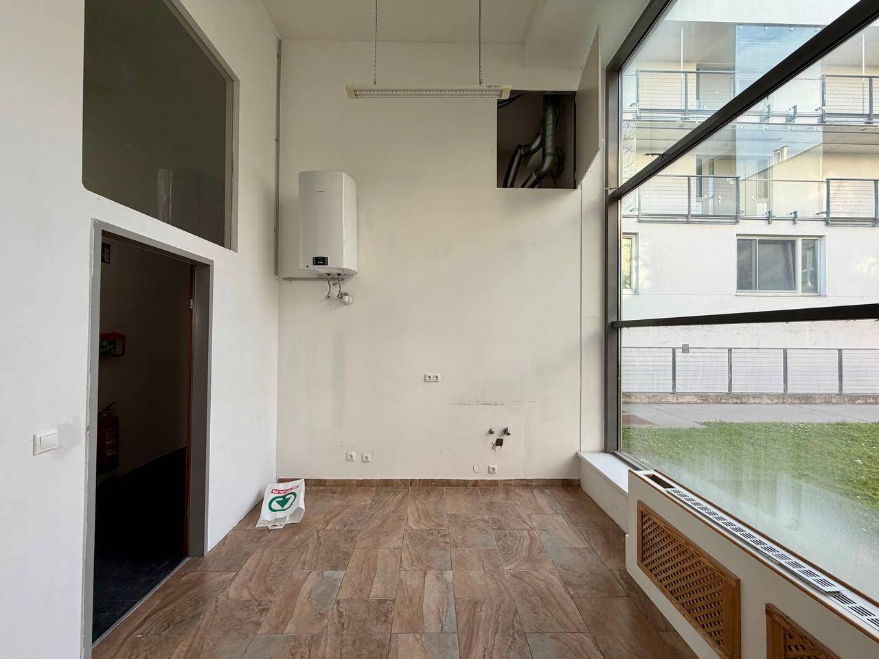 Büroraum mit großem Fenster, Warmwasserbereiter und braunem Fliesenboden.