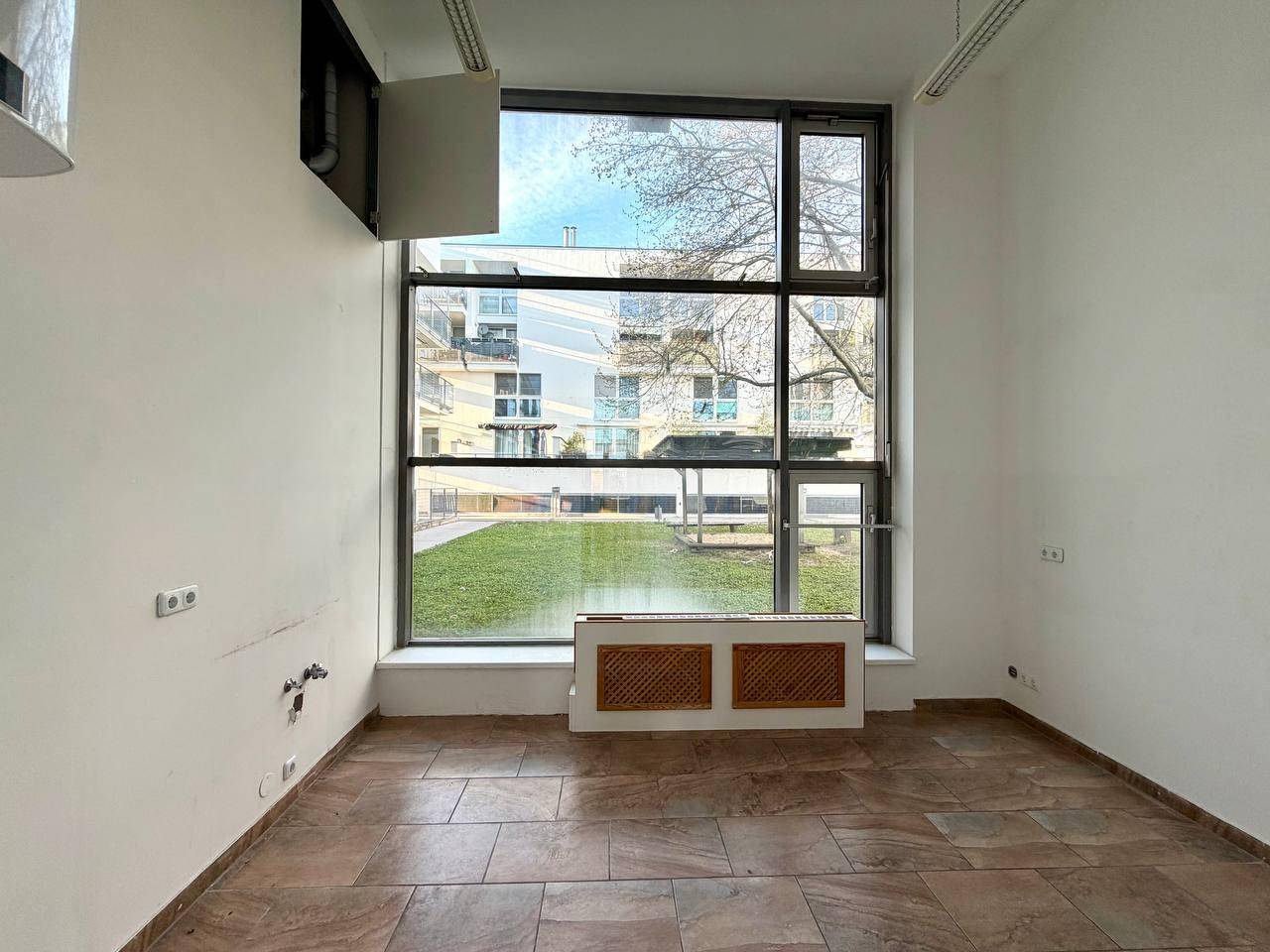 Kleiner Büroraum mit großen Fenstern, braunem Fliesenboden und Blick ins Grüne.
