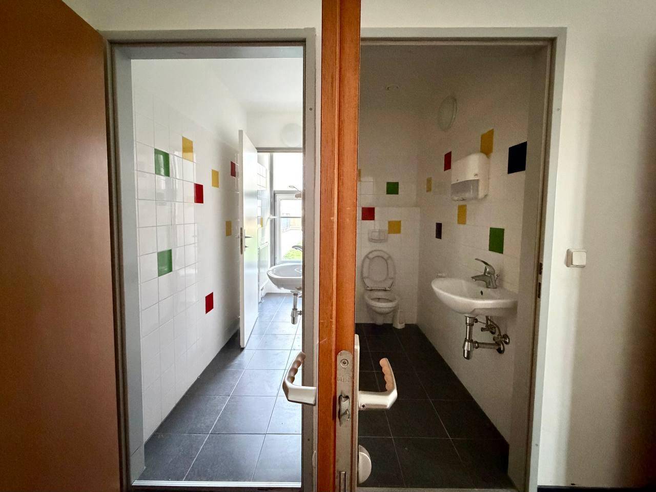 Zwei Toilettenräume mit weißen Fliesen, farbigen Akzenten und dunklem Boden.