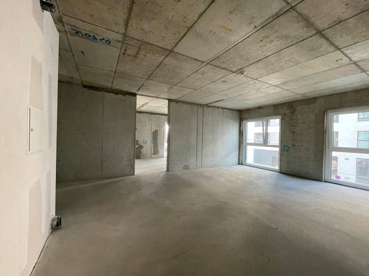 Blick in einen unfertigen Raum mit Betonwänden und -boden, gekennzeichnet durch große Fenster und Helligkeit.