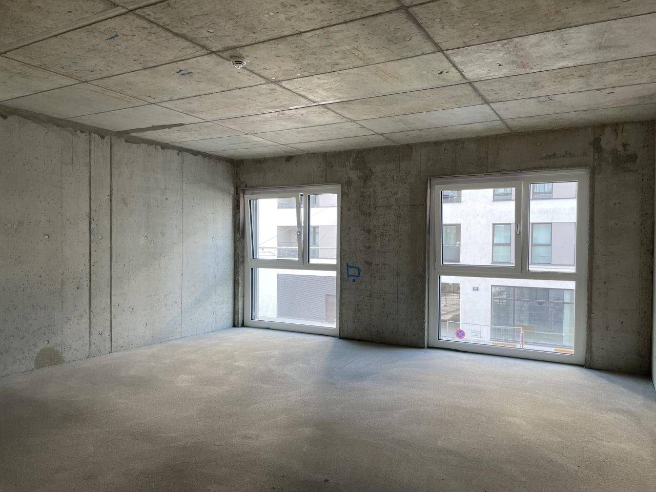 Unfertiger Innenraum mit rohen Betonwänden und -decke, großen Fenstern und viel Tageslicht.