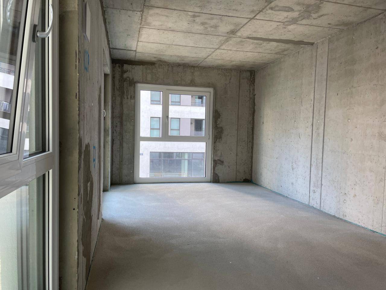 Unfertiger Innenraum mit Betonwänden und einem großen Fenster, das einen Blick auf andere Gebäude bietet.