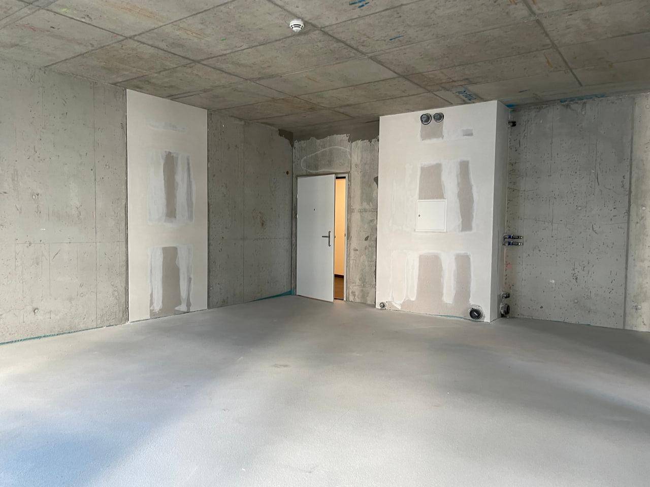 Unfertiger Raum mit Betonwänden, einer Tür und vorbereiteten Installationen für den weiteren Ausbau.