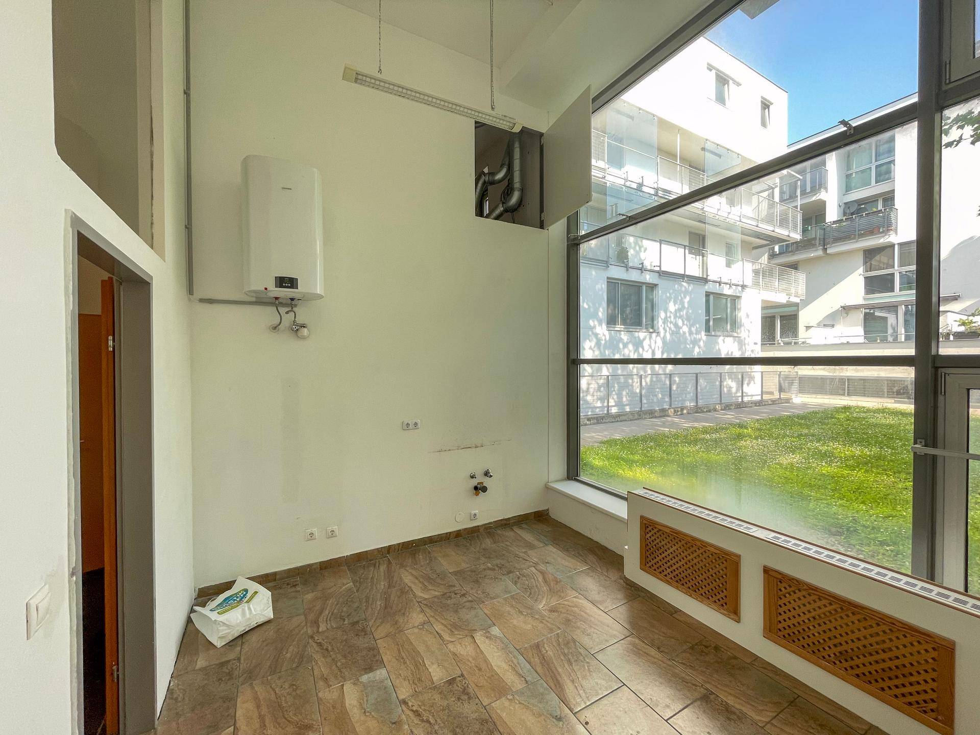 Büroraum mit großem Fenster, Warmwasserbereiter und braunem Fliesenboden.