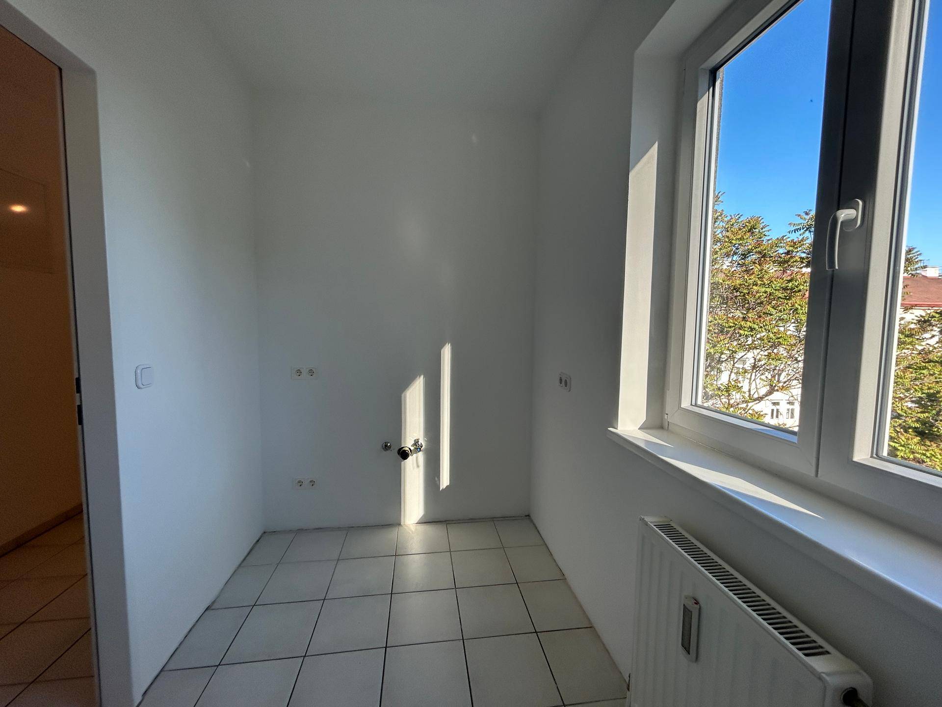 Heller Raum mit Fliesenboden, Fenster und Anschlüssen für Wasser, ideal für eine Küche.