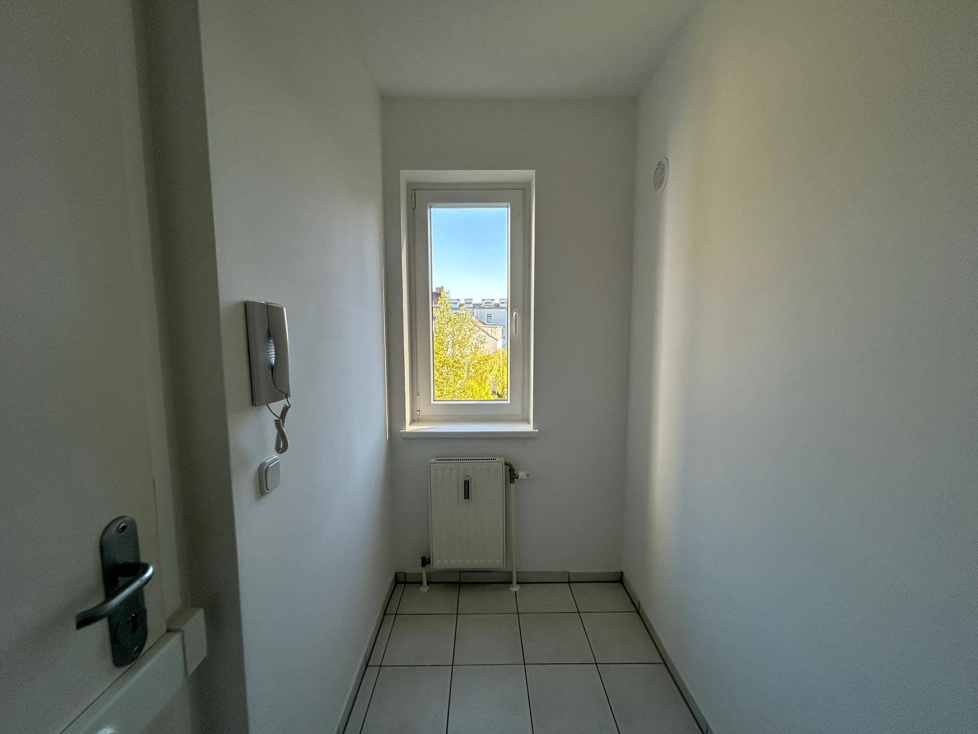 Heller Flur mit weißen Fliesen, einer Gegensprechanlage und einem Fenster mit Ausblick.