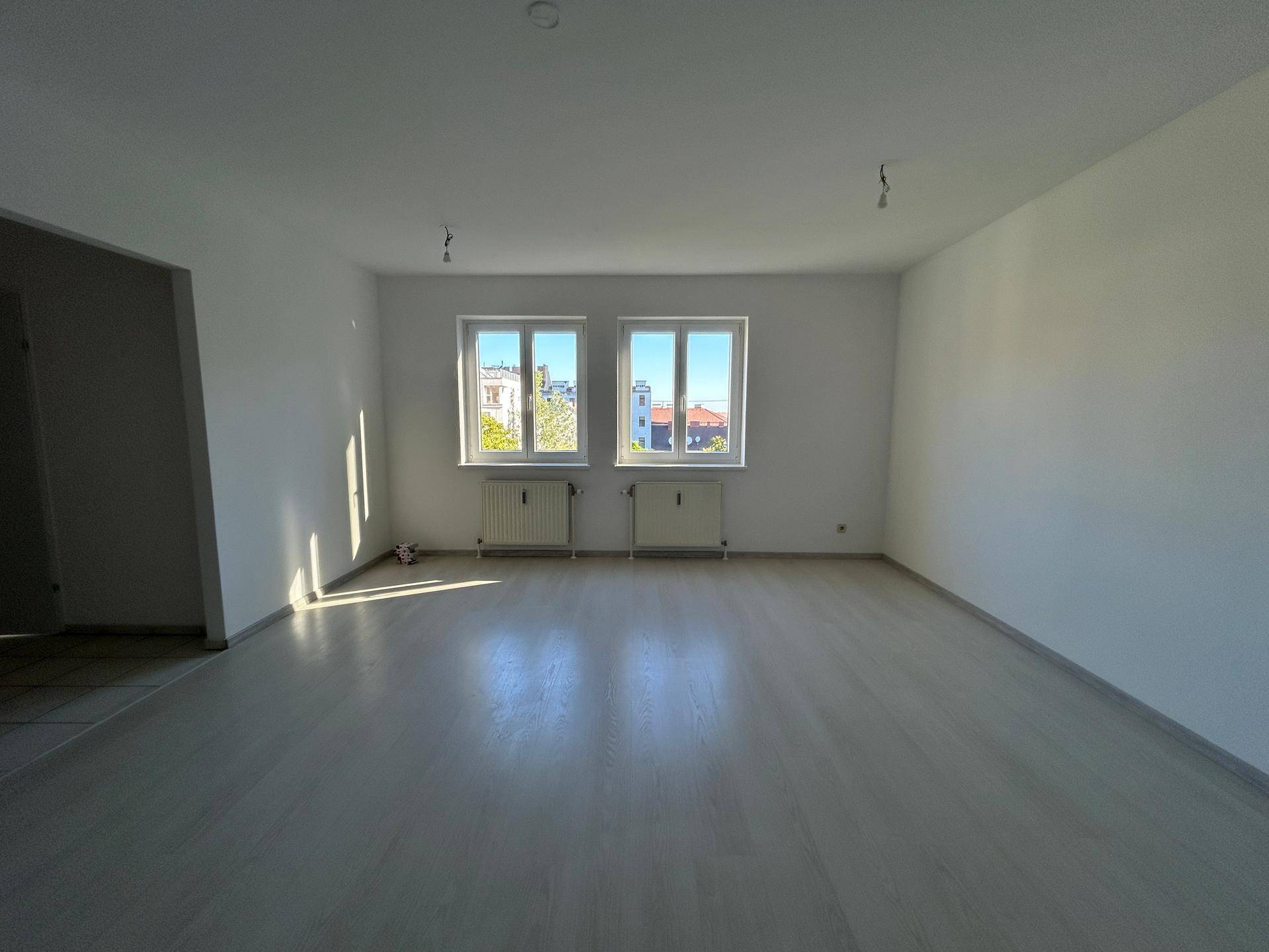 Geräumiges Wohnzimmer mit hellem Laminatboden und zwei großen Fenstern für viel Tageslicht.