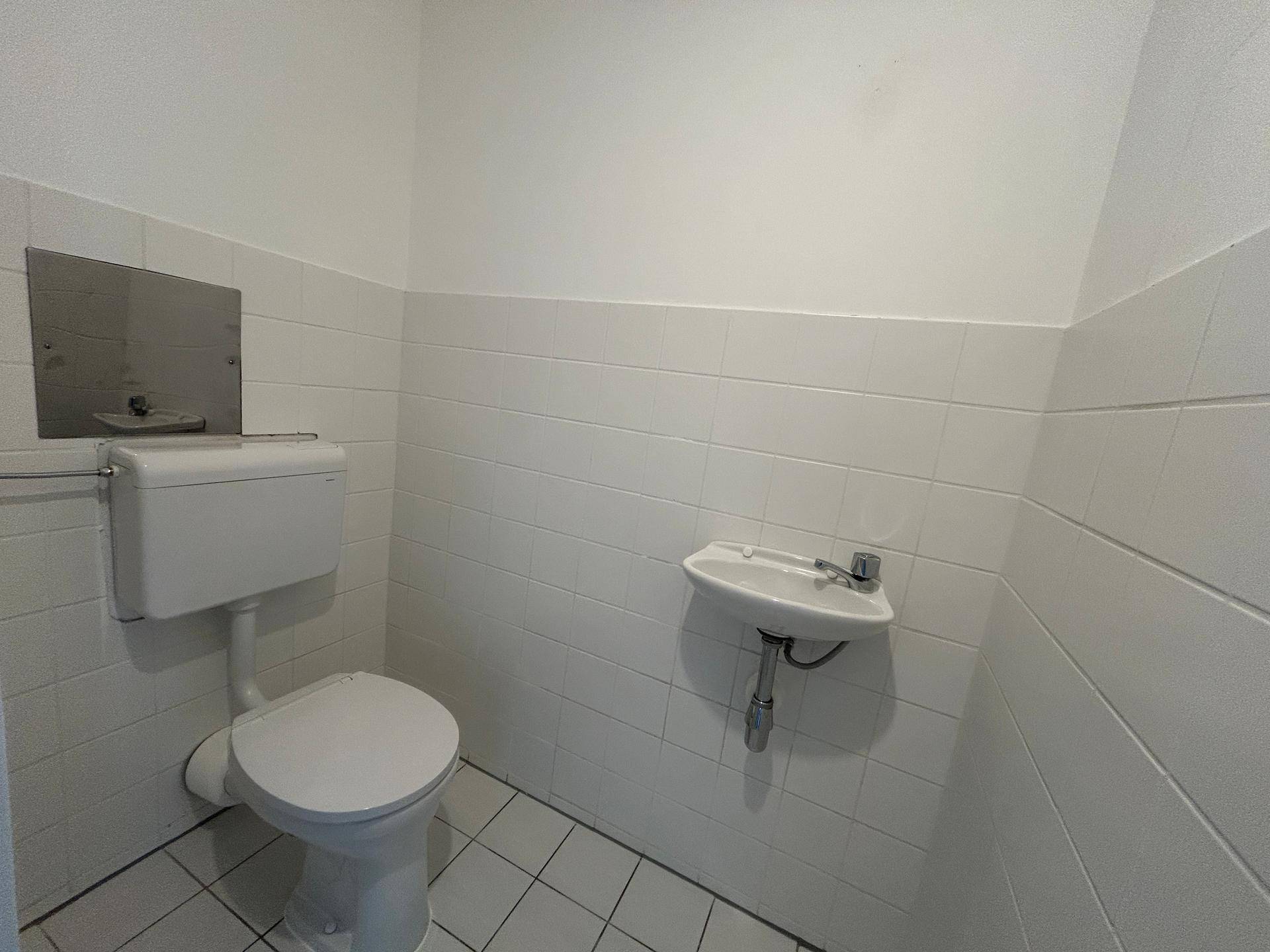 Einfache Gästetoilette mit weißen Fliesen, WC und kleinem Waschbecken.
