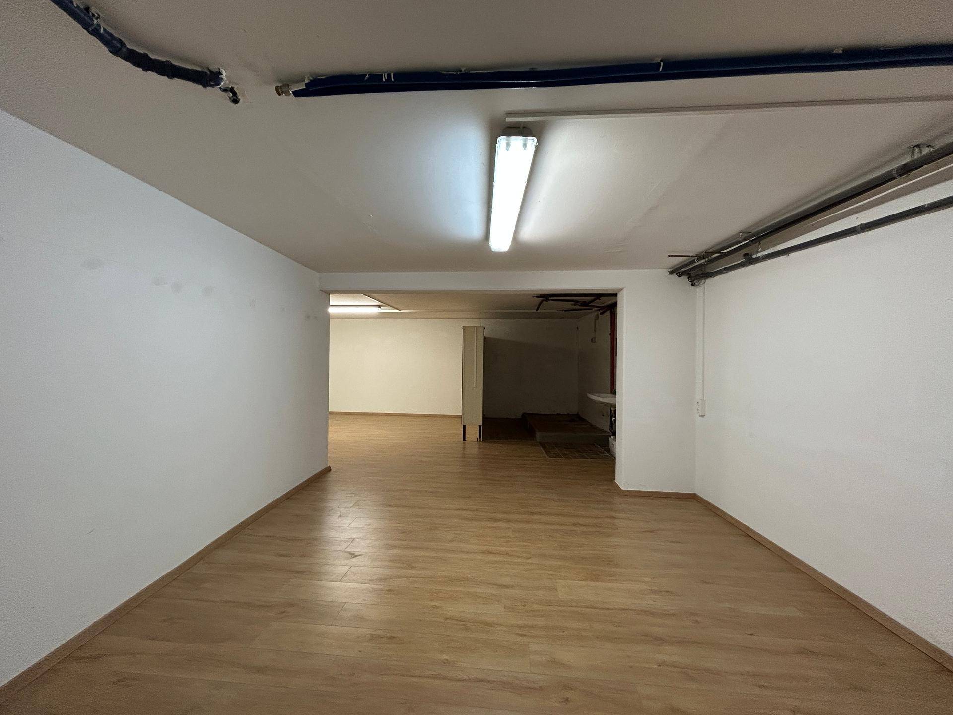Geräumiger Lagerraum mit hellem Laminatboden und sichtbaren Deckeninstallationen, gut beleuchtet.