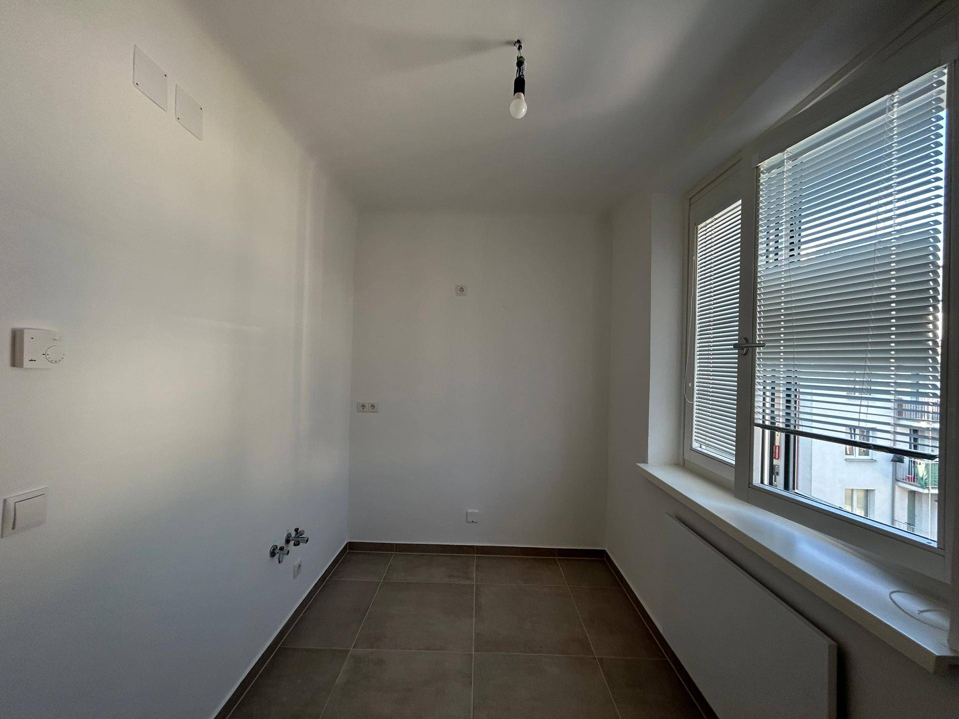 Leerer Raum mit Fliesenboden, weiß gestrichenen Wänden und einem Fenster mit Jalousien.