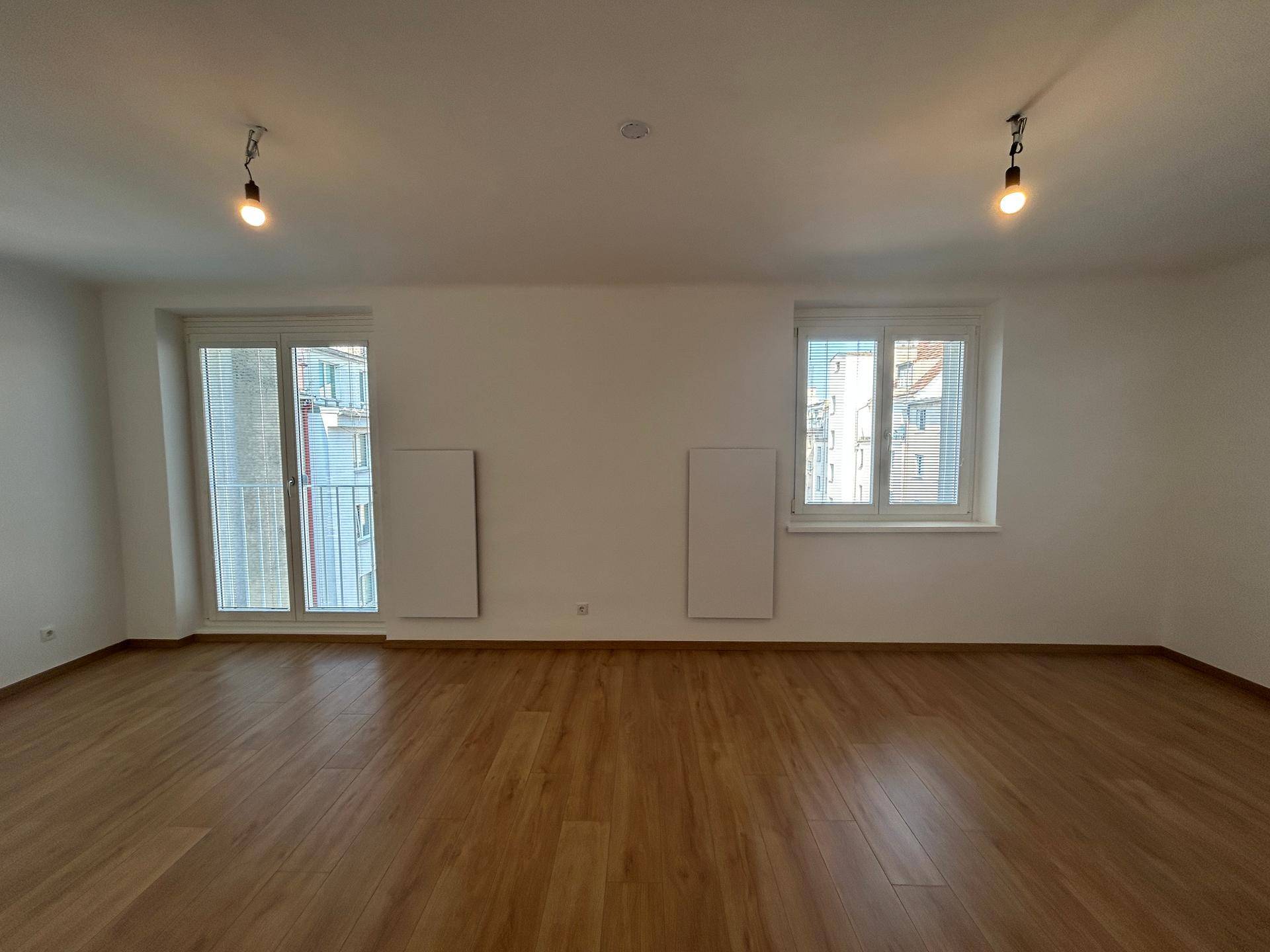 Geräumiger, heller Raum mit Holzfußboden, zwei Fenstern und Zugang zu einem französischen Balkon.