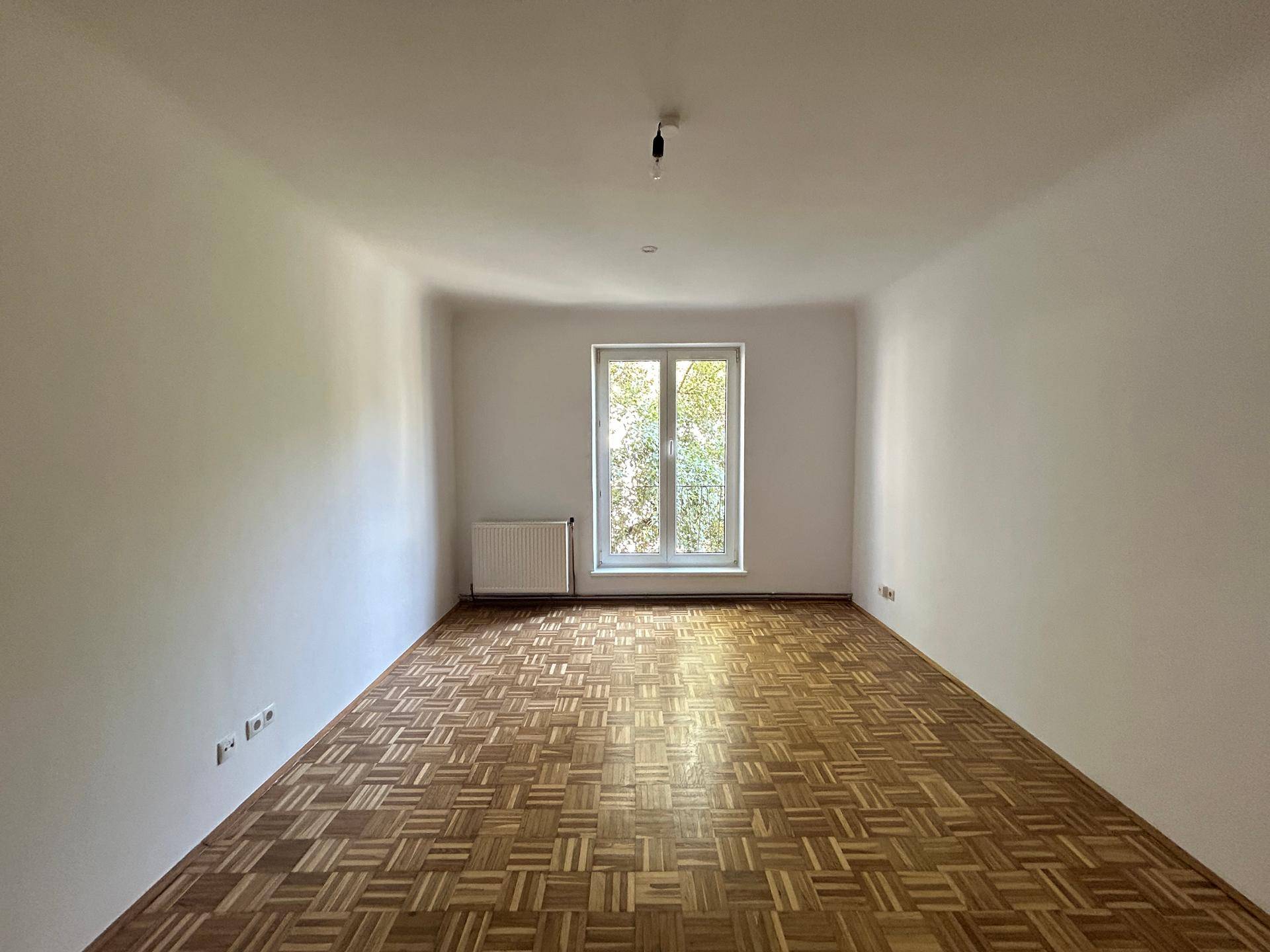 Geräumiges Wohnzimmer mit Fischgrätparkett und großem Fenster, das viel Licht hereinlässt.