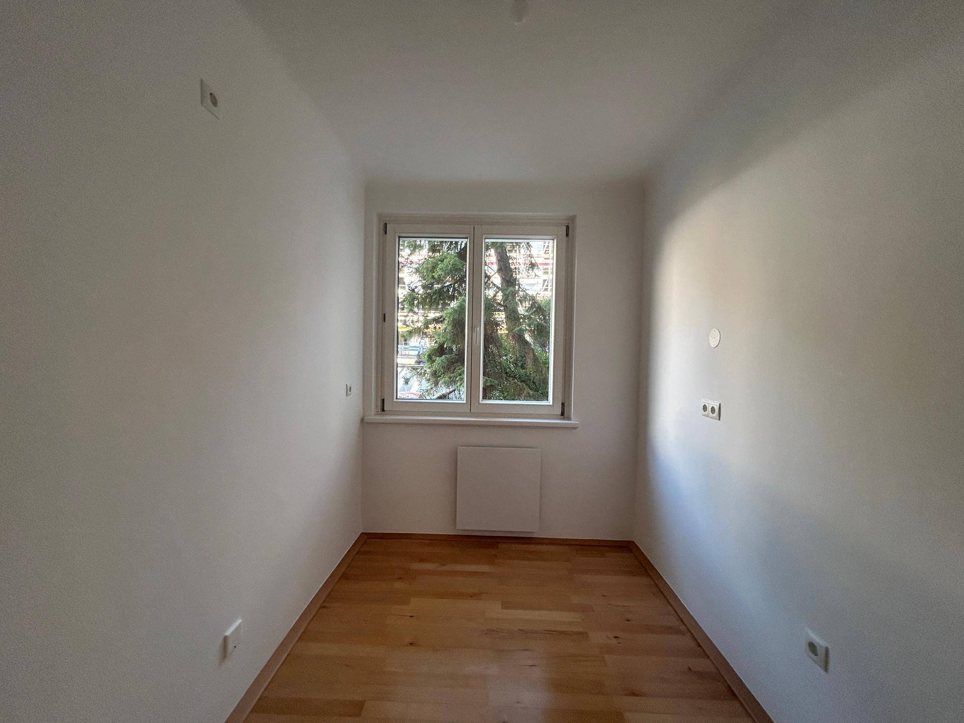 Lichtdurchflutetes Zimmer mit Holzboden und weiß gestrichenen Wänden, mit Blick ins Grüne.