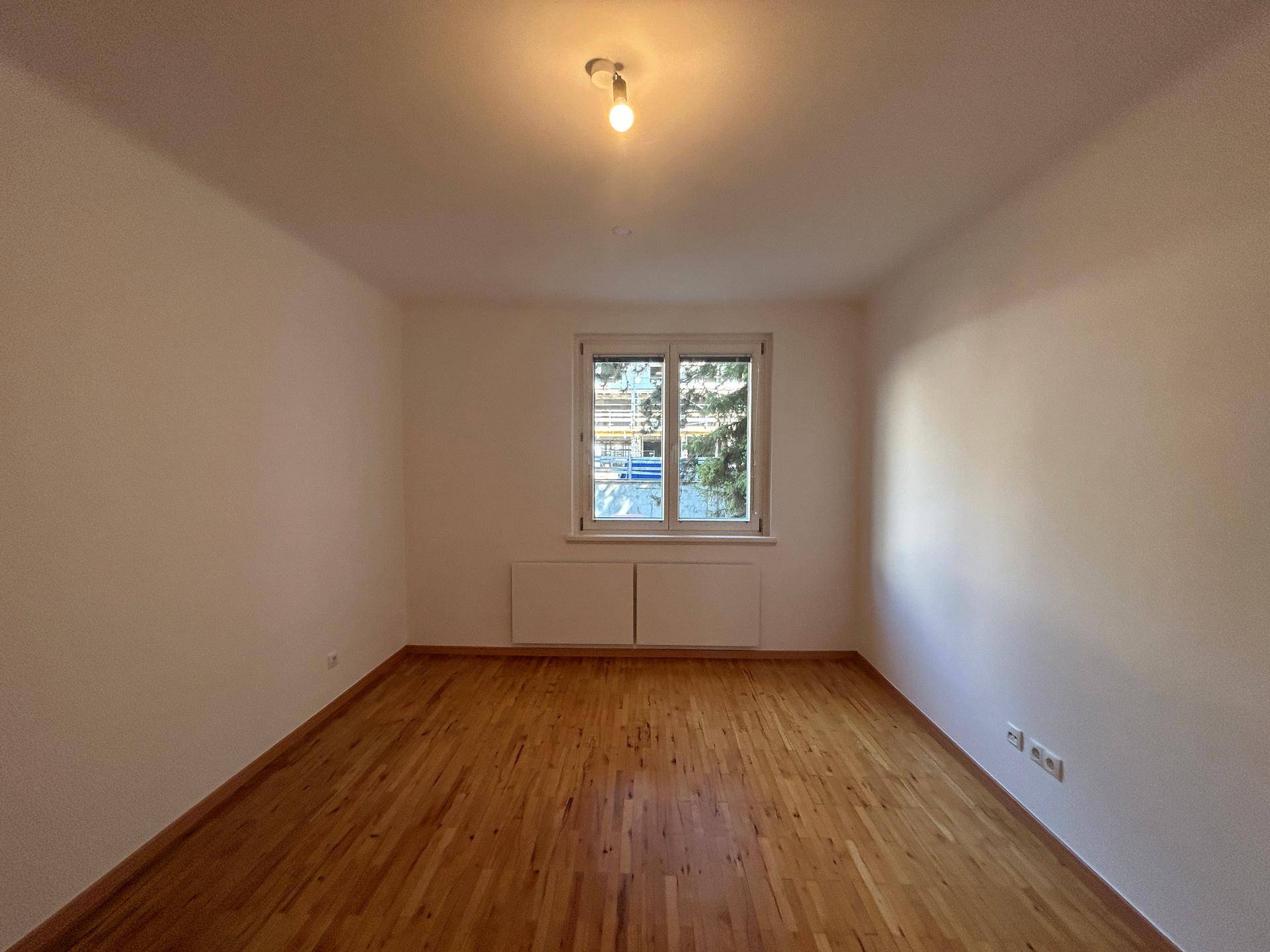 Helles Zimmer mit Holzboden und weißen Wänden, mit einem Fenster für viel Tageslicht.