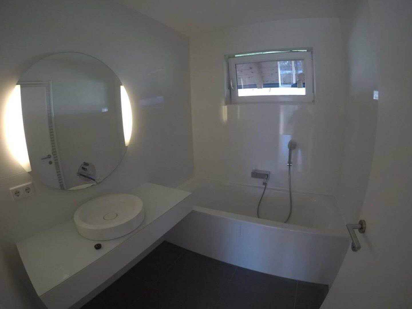 Modernes Badezimmer mit Badewanne, rundem Spiegel und dunklen Bodenfliesen.