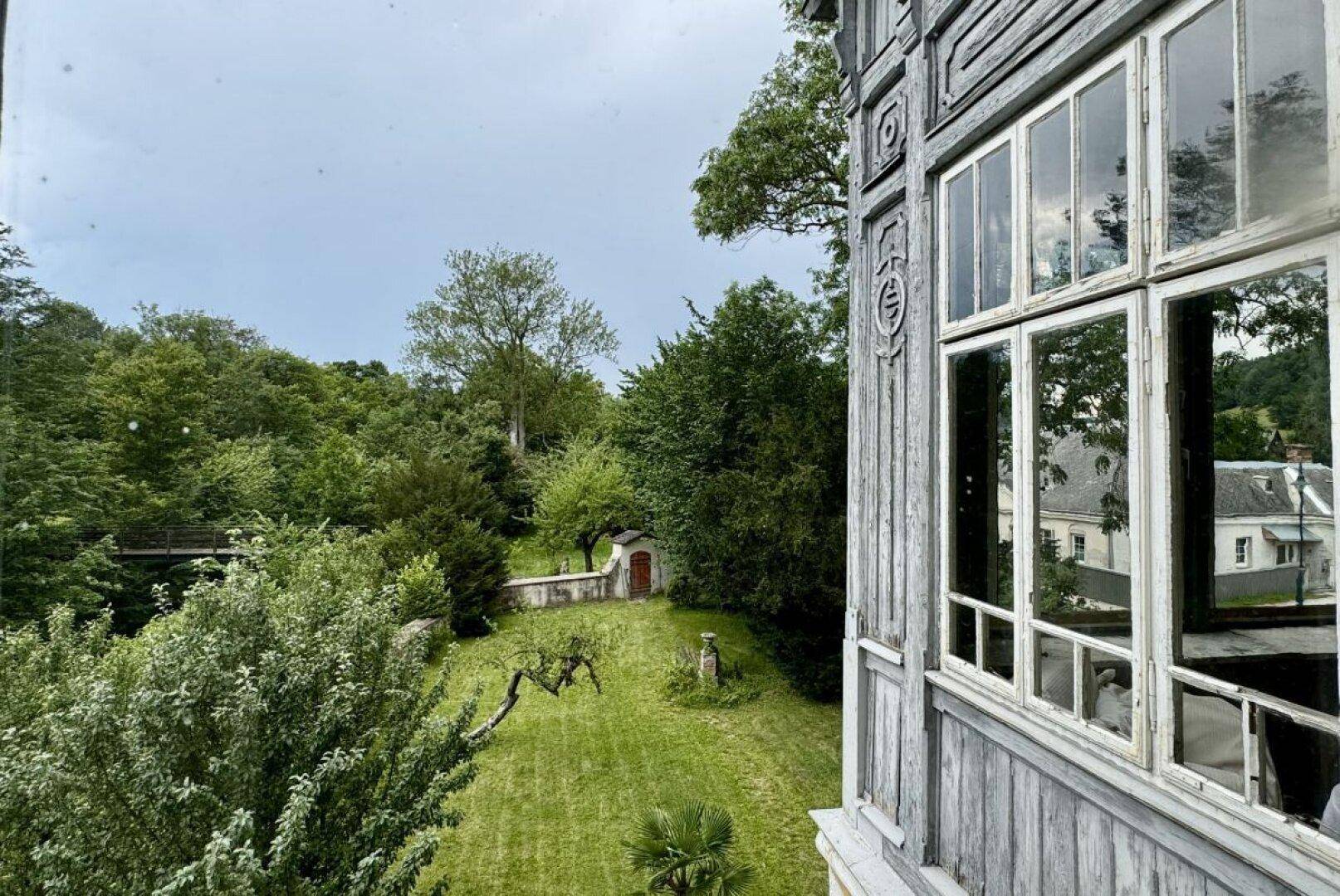Blick aus einem Fenster auf den weitläufigen Garten mit Bäumen und einem kleinen Gebäude.