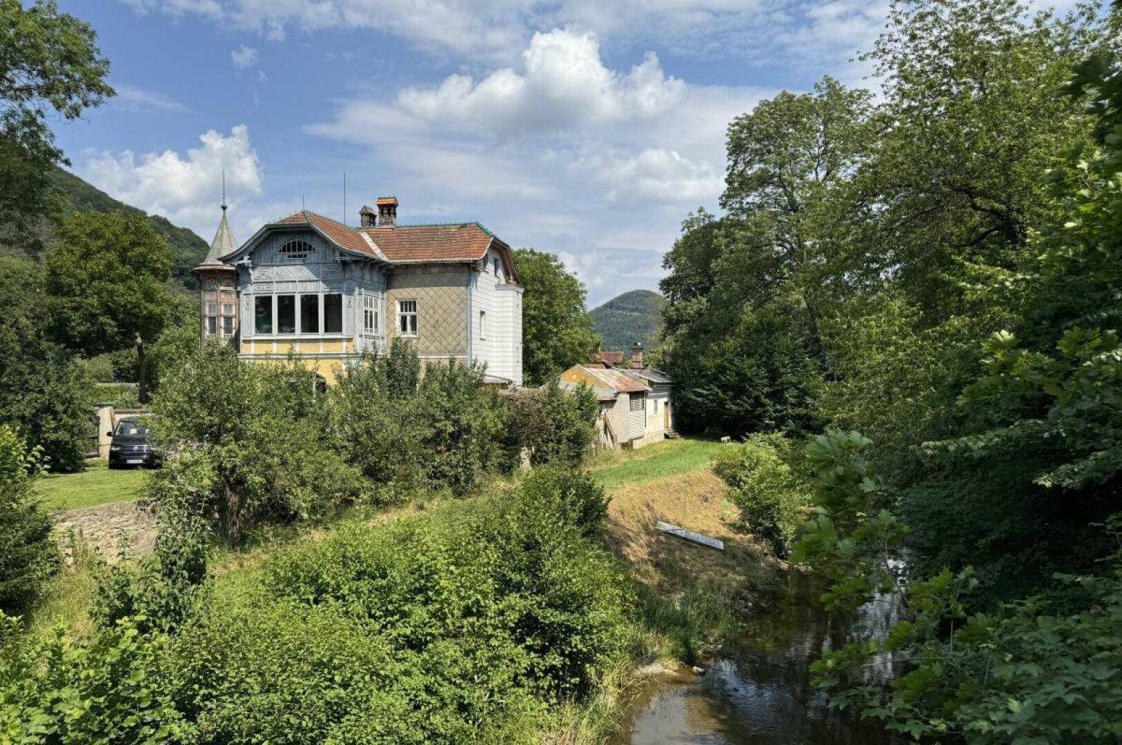 Historische Villa umgeben von üppiger Natur und einem Fluss im Vordergrund.