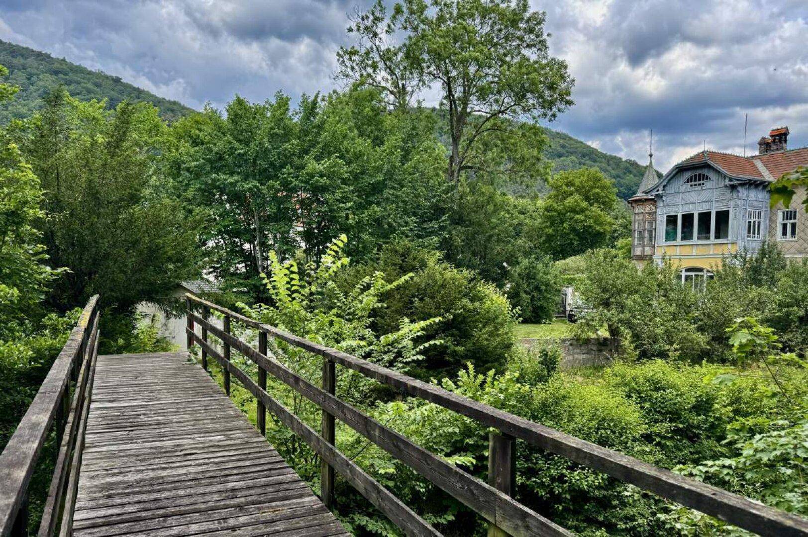 Holzbrücke führt über einen Fluss mit Blick auf die historische Villa und die umliegende Natur.