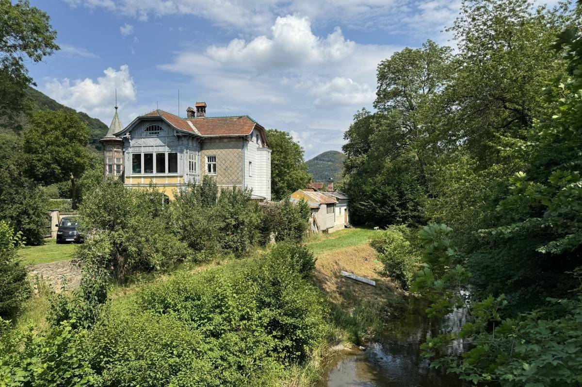 Historische Villa am Flussufer, umgeben von üppigem Grün und Bergen im Hintergrund.