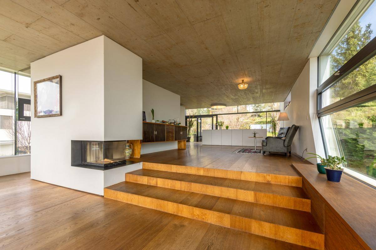 Heller Wohnbereich mit modernem Kamin, Holzstufen und Sichtbetondecke, umgeben von großen Fenstern.