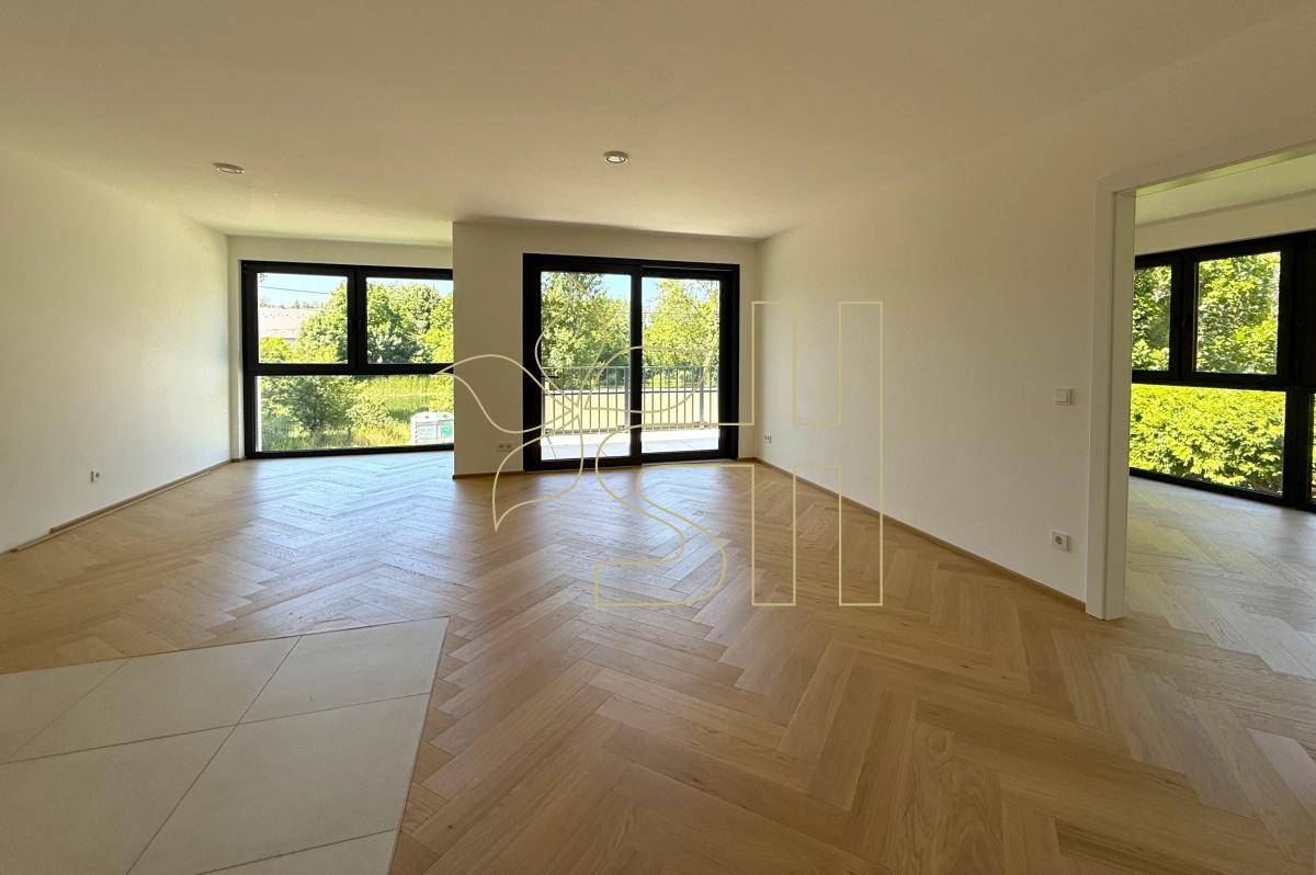 Geräumiges Wohnzimmer mit Fischgrätparkett und Zugang zu einem Balkon, umgeben von grüner Natur.