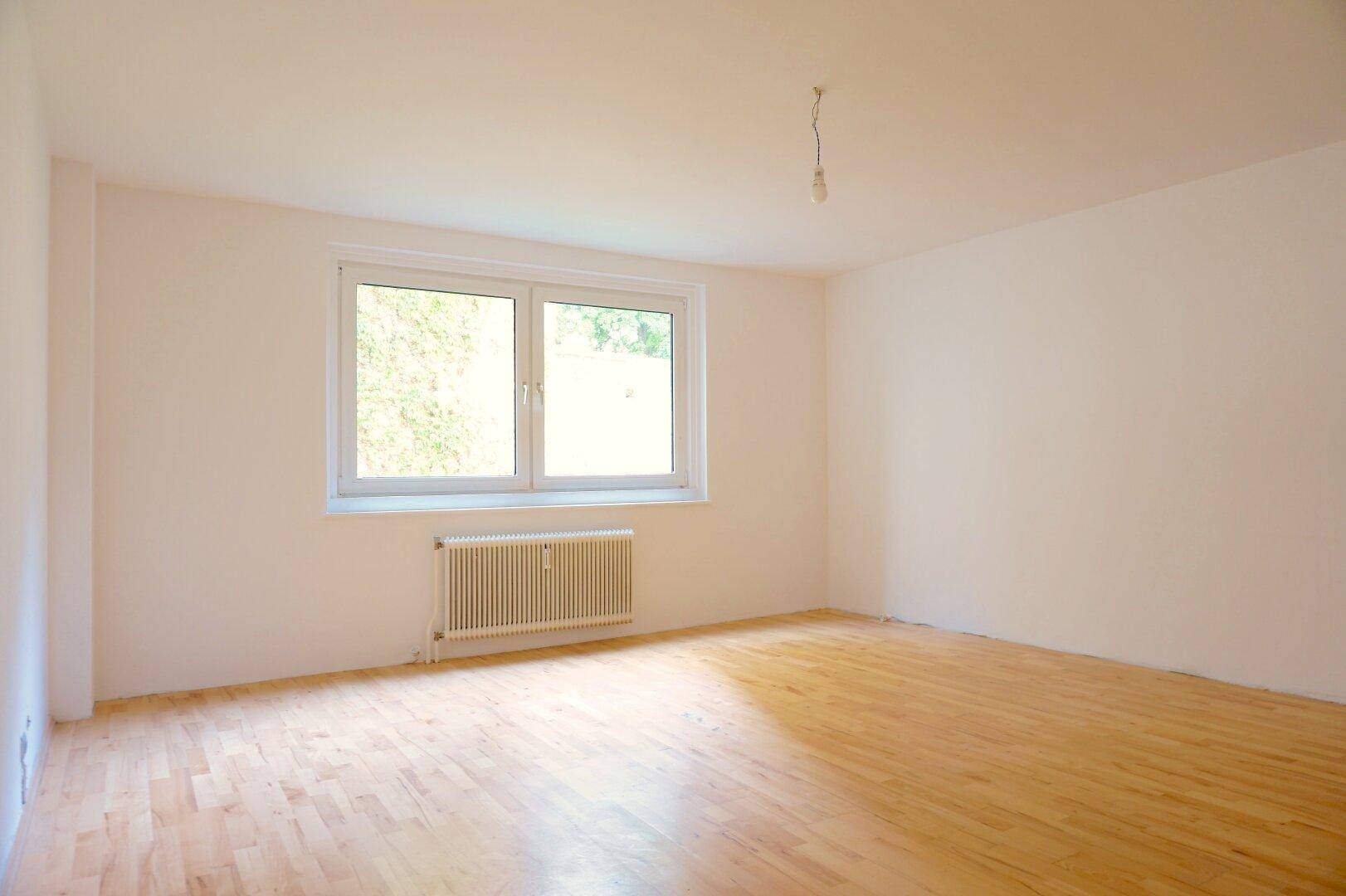 Leeres Zimmer mit hellem Holzfußboden, großem Fenster und Heizkörper, bereit zur individuellen Gestaltung.