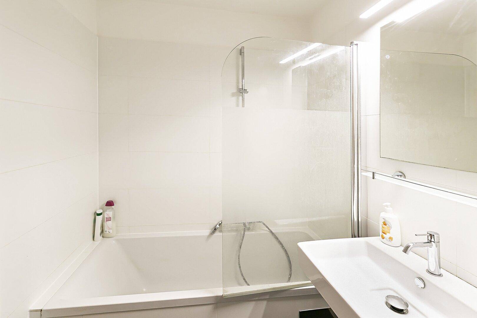 Helles Badezimmer mit weißer Badewanne, Duschabtrennung und modernem Waschbecken.
